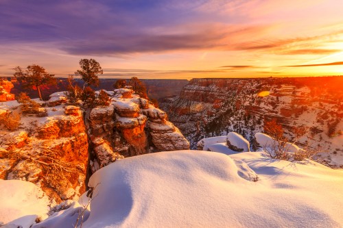 Snow-covered cliffs at the Grand Canyon during sunset, with vibrant colors illuminating the sky and landscape.