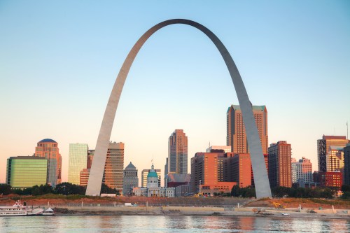 The Gateway Arch stands tall against a sunset skyline, reflecting in the water below.