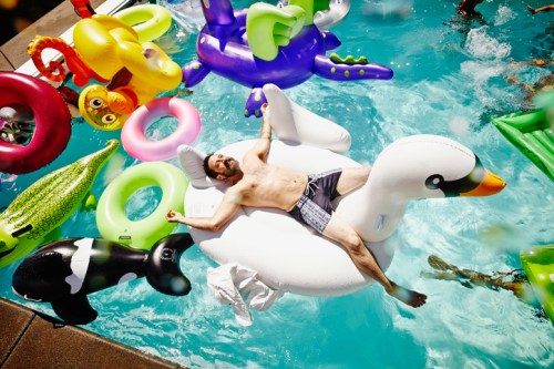 A man relaxes on a large swan float in a pool surrounded by colorful inflatable toys.