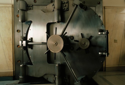 A large, metallic bank vault door with a circular locking mechanism, set against a marble wall.