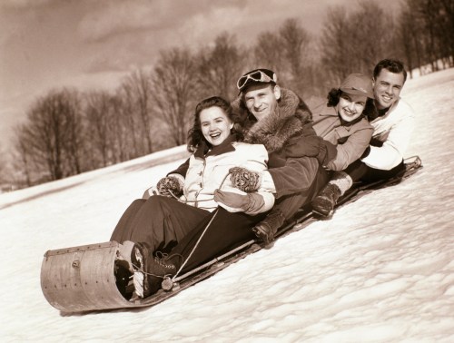 A vintage photo of four friends joyfully sledding down a snowy hill, smiling and bundled in winter clothing.