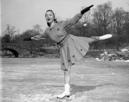 A joyful figure skater in a polka dot dress performs a graceful pose on an ice rink surrounded by trees.