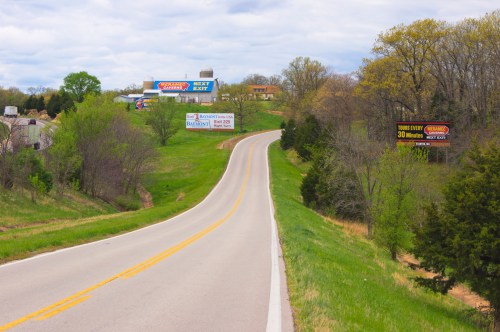 A winding road lined with green trees and billboards under a cloudy sky.