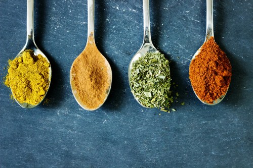 Four metal spoons on a dark surface, each filled with different spices: yellow powder, brown powder, green herbs, and red powder.