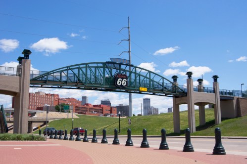 A green bridge with a Route 66 sign spans over a road, with city buildings and blue sky in the background.