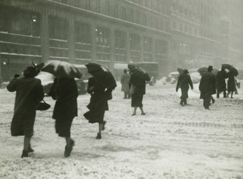 People walking in a snowstorm, holding umbrellas, with snow-covered streets and buildings in the background.
