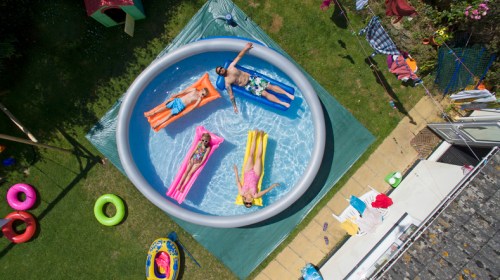 Four people relax on inflatable loungers in a circular pool, surrounded by colorful pool toys and a grassy yard.