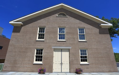 A brick building with a symmetrical facade, featuring three windows and a central double door, under a clear blue sky.