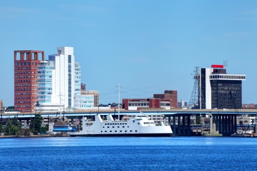 A white ferry docked on a river with a city skyline featuring modern buildings in the background under a clear blue sky.