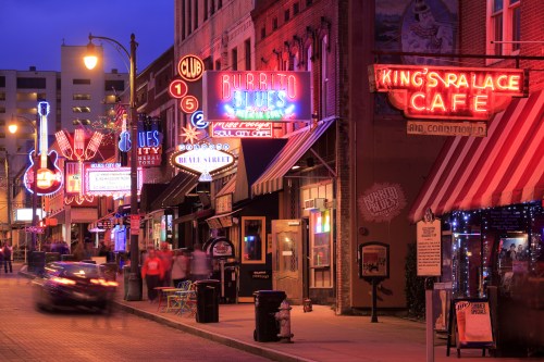 Colorful neon signs illuminate a lively street lined with restaurants and clubs at dusk.