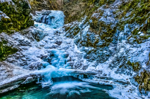 A frozen waterfall cascading over rocky terrain, surrounded by icy blue and green hues in a winter landscape.