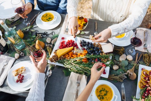 A festive table spread with a variety of fruits, cheeses, and soups, surrounded by people enjoying a meal together.