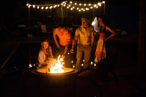 A group of four people gathered around a fire pit at night, enjoying a warm, festive atmosphere with string lights in the background.