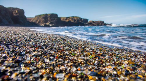 Colorful sea glass and pebbles scattered on a beach, with gentle waves lapping at the shore under a clear blue sky.