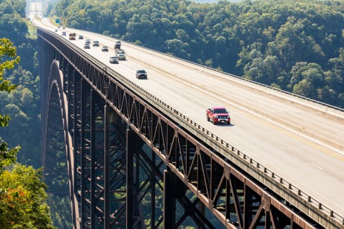 A red truck drives on a high bridge with cars, surrounded by lush green trees and hills in the background.