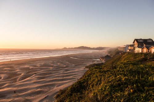 Sunset over a sandy beach with gentle waves, framed by lush greenery and coastal homes in the distance.