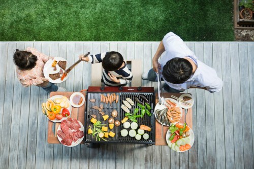A family grilling outdoors, with various meats and vegetables on the barbecue and plates of food on the table.