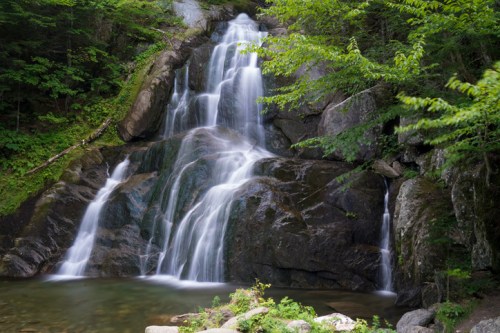A serene waterfall cascading over rocks, surrounded by lush green foliage and a calm pool at the base.
