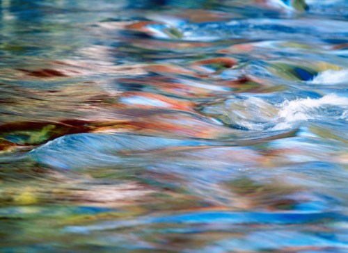 Colorful reflections ripple across the surface of a flowing river, creating a vibrant, abstract pattern.