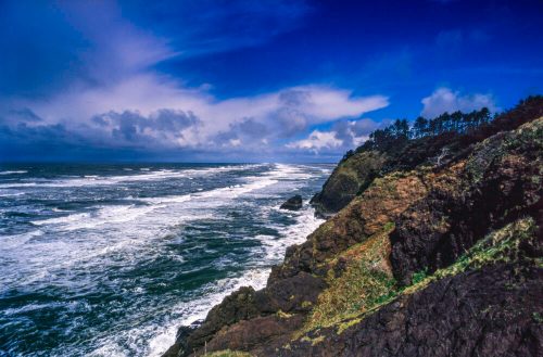 A dramatic coastal scene with waves crashing against rocky cliffs under a blue sky with scattered clouds.