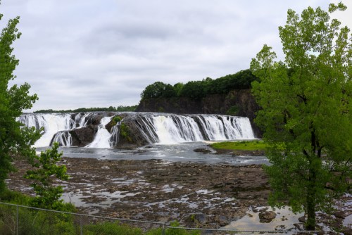 A panoramic view of a waterfall cascading over rocky cliffs, surrounded by lush green trees under a cloudy sky.