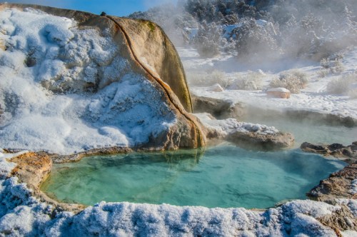 A steaming hot spring surrounded by snow-covered rocks and a winter landscape.