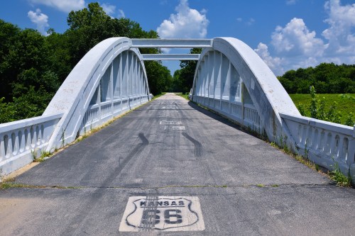 A white arch bridge spans a road, surrounded by green trees and a blue sky with fluffy clouds. Kansas Route 66 is marked on the pavement.