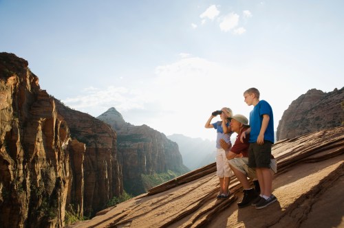 Three children explore a scenic mountain view, one using binoculars, with sunlight illuminating the landscape.