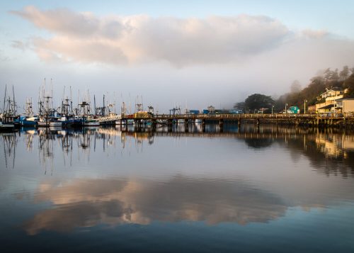 A serene harbor scene with fishing boats, a wooden pier, and foggy clouds reflecting on calm water.