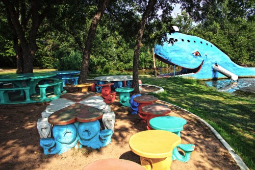 Colorful picnic tables surround a large, whimsical blue whale structure by a serene lake, shaded by trees.