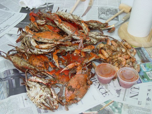 A spread of cooked crabs and shrimp on newspaper, accompanied by two containers of dipping sauce.