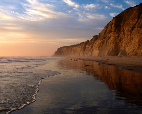 A serene beach at sunset, with cliffs in the background and gentle waves reflecting the colorful sky.