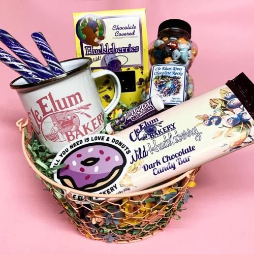 A colorful basket filled with treats: a mug, candy, stickers, and dark chocolate huckleberries on a pink background.