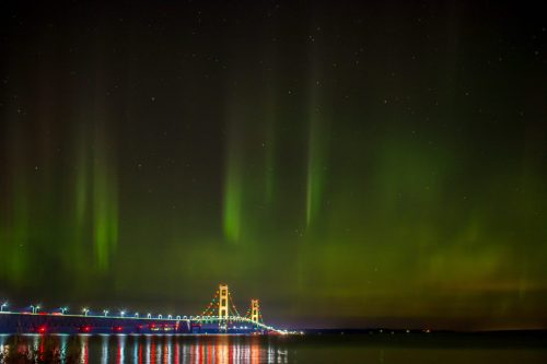 A bridge illuminated at night under vibrant green auroras and a starry sky, reflecting on calm waters below.