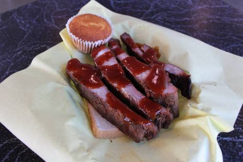 Sliced barbecue brisket with sauce on a piece of bread, accompanied by a cornbread muffin on yellow paper.