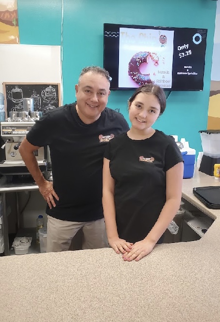 A man and a girl stand behind a counter in a cafe, smiling. A menu is displayed on a screen in the background.