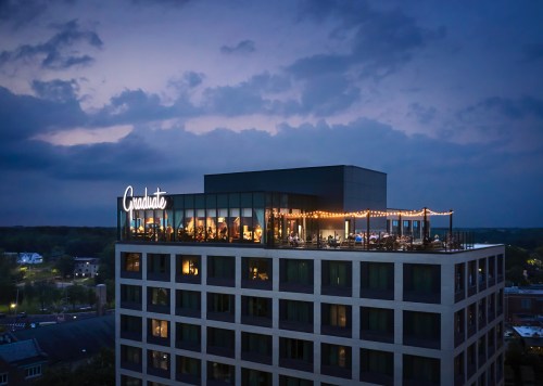 A rooftop bar with string lights on a modern building at dusk, featuring the word "Graduate" illuminated.