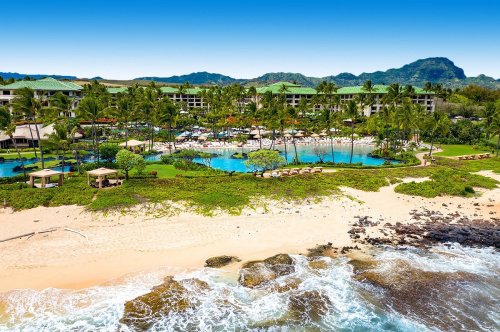 A tropical resort with a pool, palm trees, and beach, set against a backdrop of mountains and clear blue skies.