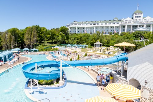 A sunny resort pool area featuring a blue water slide, lounge chairs, and a large hotel in the background.