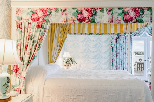 Brightly decorated bedroom with floral curtains, a white bedspread, and a view of a balcony through glass doors.