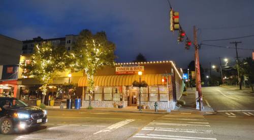 A diner and bar with a bright awning, illuminated trees, and a traffic light at dusk on a city street.