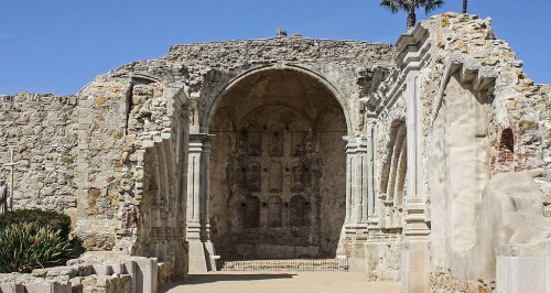 Ruins of a historic stone structure with an arched entrance and decorative elements, set against a clear blue sky.