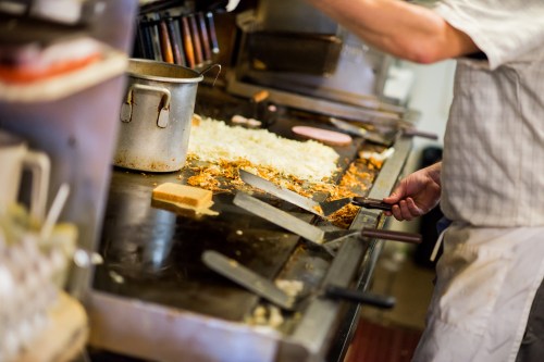 A chef cooking on a griddle with vegetables and bread, surrounded by kitchen equipment.