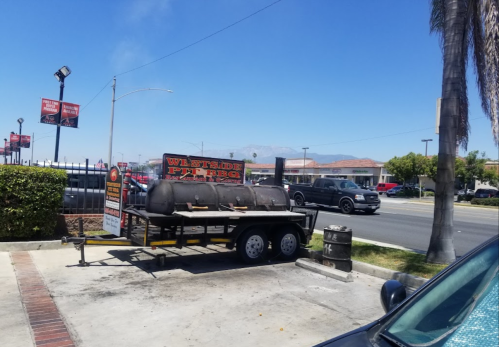 A BBQ trailer with a sign reading "Westside Pit BBQ" parked on a sunny street with palm trees and buildings in the background.