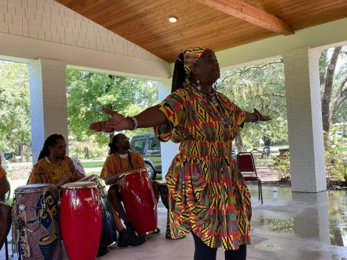 A performer in colorful attire gestures with arms outstretched, accompanied by musicians playing drums.
