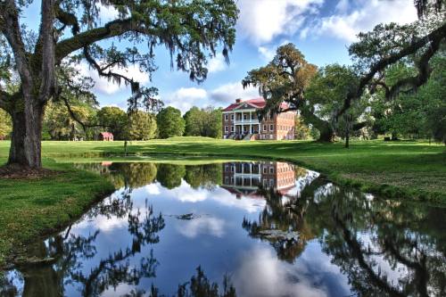 A historic building reflected in a calm pond, surrounded by lush greenery and trees draped in Spanish moss.