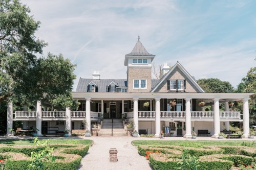 A large, elegant mansion with a tower, surrounded by gardens and a gravel pathway under a clear blue sky.