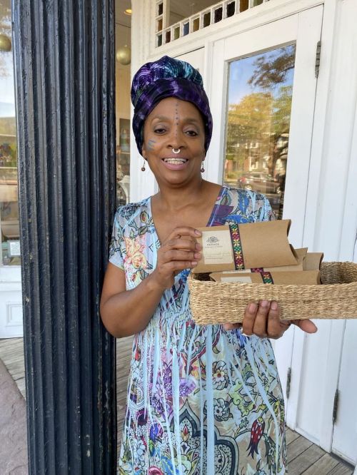 A woman in a colorful dress and headwrap holds a basket of packaged goods outside a shop.