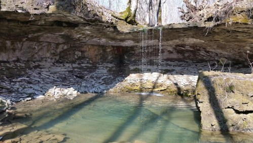 A serene natural scene featuring a small waterfall cascading into a clear pool surrounded by rocky terrain and shadows.