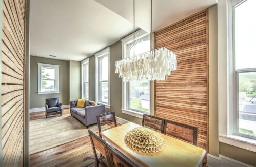A modern dining area with a wooden table, unique chandelier, and large windows, featuring a cozy seating area.